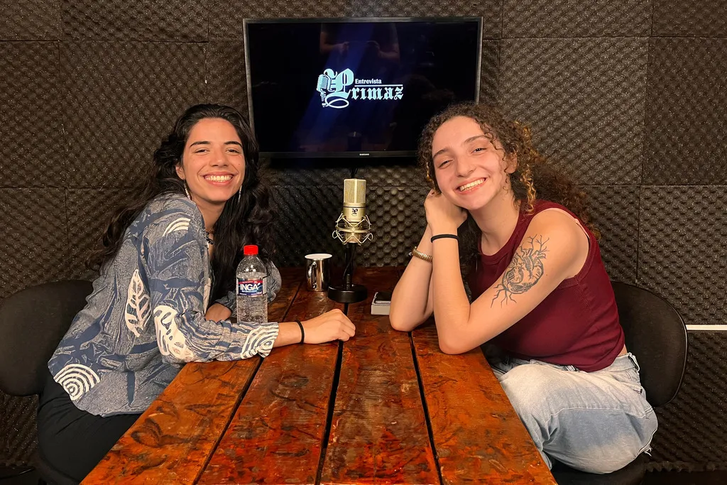 duas mulheres sorriem para  a câmera, elas estão sentadas uma de frente para a outra, uma mesa as separa, ao fundo uma tv exibe a logo da Entrevista Primaz e sobre a mesa tem um tablet, um microfone e algumas canecas