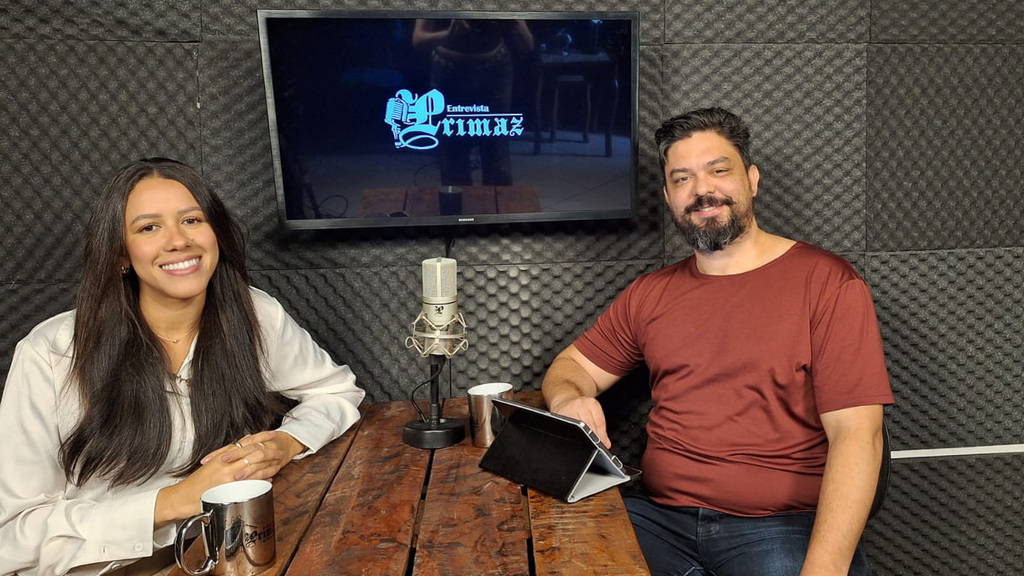 A imagem apresenta um cenário de estúdio de podcast com duas pessoas sentadas à volta de uma mesa de madeira rústica, posicionadas em frente a uma parede revestida com espuma acústica cinza-escura em formato de caixa de ovo. As Pessoas À esquerda: A nutricionista Rayane Souza. Ela é uma mulher jovem de pele clara e longos cabelos castanhos escuros. Está sorrindo para a câmera, veste uma blusa social branca de mangas longas e calças bege. Seus braços estão apoiados na mesa, e ela segura uma caneca metálica. À direita: O jornalista Lui Pereira. Ele é um homem de pele clara, com barba e bigode grisalhos e cabelos curtos escuros penteados para trás. Ele sorri de forma amigável, veste uma camiseta bordô e calças jeans escuras. Sua mão esquerda repousa sobre um tablet posicionado na mesa. O Cenário e Equipamentos Centro da Mesa: Um microfone condensador profissional de cor prateada está fixado em um suporte de mesa entre os dois participantes. Sobre a Mesa: Além da caneca de Rayane e do tablet de Lui, há um smartphone branco deitado próximo à nutricionista e uma segunda caneca metálica perto do jornalista. A mesa é feita de tábuas de madeira robustas com marcas de desgaste natural. Fundo: Centralizada na parede atrás deles, há uma televisão de tela plana, onde é possível notar a logo do programa Entrevista Primaz.