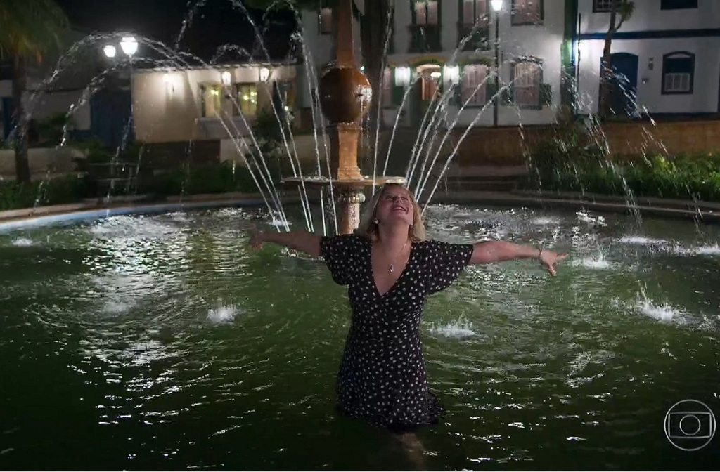 Mulher de vestido preto com estampa clara está de pé dentro de uma fonte iluminada à noite, com água até a altura das coxas. Ela abre os braços e inclina a cabeça para trás, sorrindo, enquanto jatos de água formam arcos ao redor. Ao fundo, há edificações com janelas iluminadas e postes de luz, criando uma atmosfera noturna acolhedora.