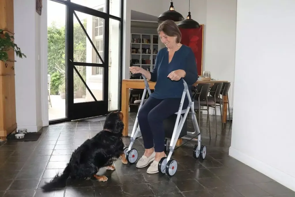 Uma mulher está sentada em uma cadeira de rodas em frente a um cachorro preto, ambos estão dentro de uma casa, ao fundo conseguimos ver uma porta de vidro entreaberta e uma mesa de jantar, além de uma estante de livros