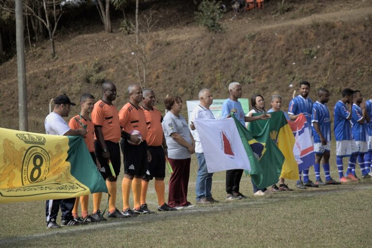 No campo do União, em Passagem de Mariana, Minas Gerais, autoridades, árbitros e atletas estão perfilados para a abertura do Campeonato Amador de Futebol, segurando as bandeiras das equipes do 8 de Dezembro e São Caetanense, e também do Brasil e de Minas Gerais