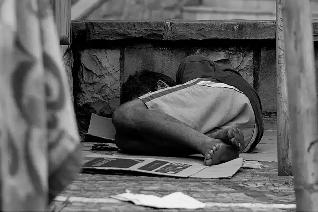 Em um canto de rua, sobre um papelão colocado no chão, uma pessoa dorme, protegendo-se dos ciclos do clima