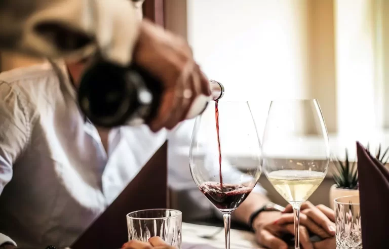 Mão de um garçom segurando garrafa e servindo vinho em uma taça, ilustrando a crônica Reaprendendo a ver gente reunida