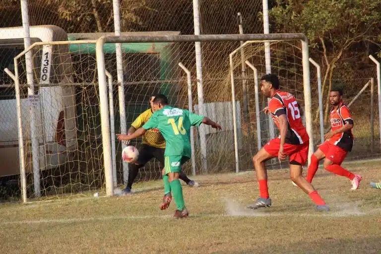 Em partida do Campeonato Amador de Mariana, jogador do 8 de Dezembro, com uniforme verde e amarelo, prepara-se para chutar diante do goleiro do Bandeirantes. Dois jogadores de uniforme vermelho tentam impedir a conclusão da jogada.