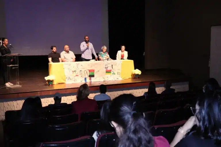 Ao fundo, sobre o palco, mesa de abertura do evento sobre Setmebro Amarelo, com quatro pessoas sentadas e, entre elas, o prefeito interino, Ronaldo Bento, de pé, fala à plateia posicionada em primeiro plano da imagem, sob luzes apagadas