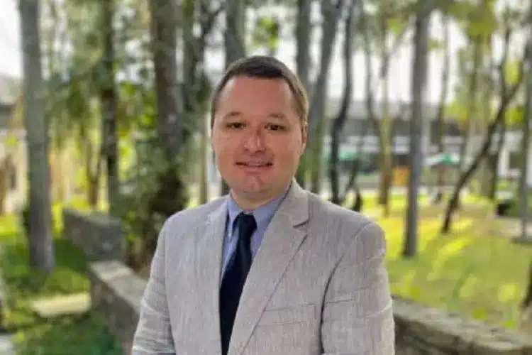 Joshua Scacco, professor da Universidade do Texas, de terno cinza claro, camisa azul e gravata preta, em foto diante de um gramado e algumas árvores