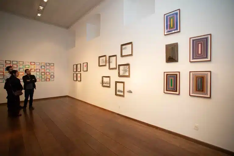 Sala do anexo do Museu da Inconfidência com quadros de diferentes artistas nas paredes. Ao lado esquerdo, três espectadores contemplando as obras.