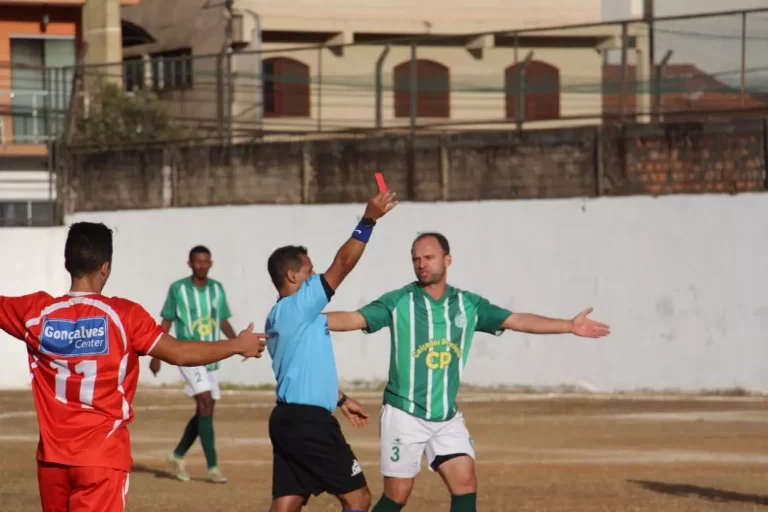 Juiz de futebol exibe cartão vermelho para jogador do Guarany, de calção branco e camisa em verde e branco, na partida contra o Olimpic, pelo Campeonato de Mariana