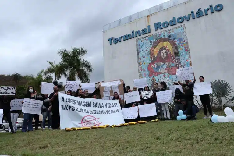 Manifestantes da área de Enfermagem seguram cartazes diante de um painel de ladrilhos colorido, no Terminal Rodoviário de Mariana