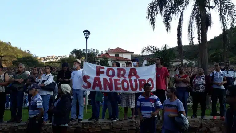 Manifestantes reunidos na praça defronte à Prefeitura de Ouro Preto portam faixa com os dizeres "Fora Saneouro"