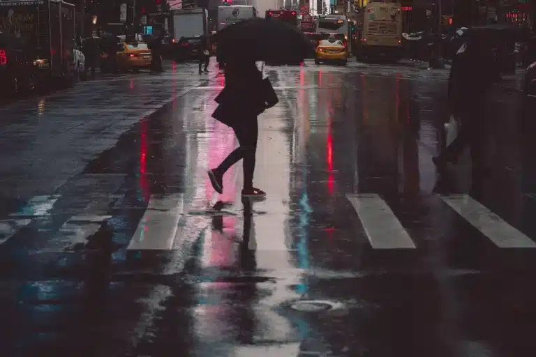 Sob chuva, mulher atravessa a faixa de pedestre á noite, protegendo-se com uma sombrinha