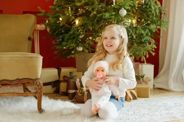 Menina, diante de uma árvore de Natal, sorri e brinca com uma boneca, seu presente de Natal