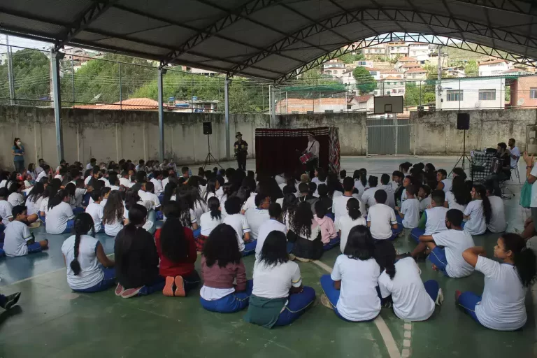 Crianças assentadas no chão de uma quadra, assistem apresentação de uma das ações educativas promovida pelo Circovolante