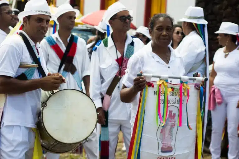 Pessoas em roupas típicas brancas, com uma mulher à frente, portando um estandarte, participam de cortejo da Festa do Reinado