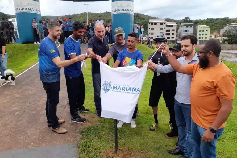 Pessoas levantam um pano branco, onde se lê: "PREFEITURA DE MARIANA, MAIS TRABALHO, NOVAS CONQUISTAS", que cobre a placa de inauguração da pista de pump track